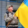 Profile photo of Ian Kaznaski, a man in camouflage attire holding a yellow and blue flag.